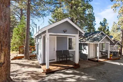 a small white cabin with a porch in the woods at Baby Bear - A delightful studio style property in the perfect central location! in Big Bear Lake