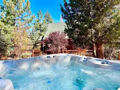 a hot tub in a yard with trees and a house at Kubo Del Sol - DREAM LOCATION WITH HOT TUB! Beautiful, light and bright log cabin! in Big Bear Lake