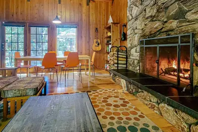 a living room with a stone fireplace and a table at Quiet Wooded Retreat in Lake Arrowhead