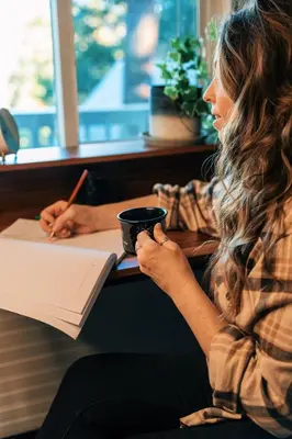 a woman sitting at a table holding a cup and writing at Little Wolf Lodge Modern Cabin at Lake Gregory in Crestline